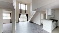 This two-story residence makes an immediate impression with its dramatic double-height ceiling in the main living area, creating a sense of spaciousness and grandeur. The open-concept living space is bathed in natural light, courtesy of a stacked pair of large windows positioned high on the wall, complemented by a sliding glass door that leads to a private balcony with outdoor views. Dark curtains frame each window, offering contrast against the crisp white walls throughout.

The living area features smooth grey hardwood-style flooring that extends throughout the main level, lending the space a clean, contemporary aesthetic. A white staircase with metal railings rises along one wall, providing both functional access to the upper floor and serving as a subtle architectural feature. Adjacent to the living space, a partial kitchen counter is visible, hinting at an open kitchen layout that connects seamlessly to the main living area. The white cabinetry and neutral finishes in the kitchen maintain the cohesive, modern palette found throughout. Overall, the space combines clean lines, high ceilings, and an abundance of natural light for a bright and airy atmosphere.