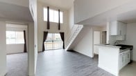 This two-story residence makes an immediate impression with its dramatic double-height ceiling in the main living area, creating a sense of spaciousness and grandeur. The open-concept living space is bathed in natural light, courtesy of a stacked pair of large windows positioned high on the wall, complemented by a sliding glass door that leads to a private balcony with outdoor views. Dark curtains frame each window, offering contrast against the crisp white walls throughout.

The living area features smooth grey hardwood-style flooring that extends throughout the main level, lending the space a clean, contemporary aesthetic. A white staircase with metal railings rises along one wall, providing both functional access to the upper floor and serving as a subtle architectural feature. Adjacent to the living space, a partial kitchen counter is visible, hinting at an open kitchen layout that connects seamlessly to the main living area. The white cabinetry and neutral finishes in the kitchen maintain the cohesive, modern palette found throughout. Overall, the space combines clean lines, high ceilings, and an abundance of natural light for a bright and airy atmosphere.