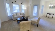 This single-story residence centers around a spacious, open-concept living area distinguished by notably high ceilings that amplify the sense of airiness throughout. The walls are finished in soft, neutral tones that complement the plush, light-colored carpeting underfoot, creating a cohesive and calming aesthetic.

The living area is well-lit by multiple windows dressed with horizontal blinds, allowing an abundance of natural light to fill the room. A glass-paneled entry door further enhances the brightness, providing an inviting transition between interior and exterior. An arc floor lamp adds a modern, stylish accent alongside the natural light sources.

The living space is thoughtfully arranged with a cushioned sofa accented by decorative pillows, a pair of patterned accent chairs in a gold geometric print, and a dark oval coffee table serving as the room's focal point. Decorative wall accents and framed artwork add personality to the otherwise understated walls.

Adjacent to the living area, a defined dining space is visible, featuring a compact console table beneath a pendant light fixture, further demonstrating the home's cohesive and functional layout. Overall, the space conveys a clean, modern, and welcoming ambiance.