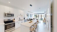 This contemporary 1-bedroom, 1-bathroom unit showcases a thoughtfully designed open-concept layout with a clean, modern aesthetic defined by an all-white palette and light flooring throughout. The kitchen is a standout feature, offering full-height white cabinetry with matte black hardware, a white tile backsplash, stainless steel appliances including a French door refrigerator, electric range, and built-in microwave, as well as a generous island with quartz countertops and bar seating — ideal for both cooking and casual dining. A sleek linear pendant light anchors the space above. The living room flows seamlessly from the kitchen and is bathed in natural light through expansive floor-to-ceiling windows, offering views of the surrounding greenery. A wall-mounted mini-split system ensures year-round comfort. The bedroom is a calm, well-proportioned retreat with its own mini-split unit and direct sightlines to the main living area. The bathroom features a frameless glass walk-in shower and built-in open shelving for convenient storage. A dedicated laundry area with a full-size washer and dryer, topped by overhead cabinetry, adds everyday practicality. This unit combines efficient use of space with high-end finishes for a refined, move-in-ready living experience.