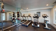 This fitness center is a dedicated, single-level workout facility designed to accommodate a well-rounded exercise regimen. The space features durable rubber flooring throughout, providing a safe and resilient surface ideal for a variety of physical activities. Recessed lighting illuminates the room evenly, creating a bright and energizing atmosphere conducive to focused training sessions.

The cardio area is well-appointed with multiple treadmills and elliptical machines, as well as a stationary bike, offering users a range of options for cardiovascular exercise. A cable machine provides versatile strength training capabilities, while a medicine ball rack stocked with various weighted balls supports functional and core training. An adjustable weight bench rounds out the strength training options.

A defining architectural feature of the space is the large floor-to-ceiling mirror panels mounted along one wall, which visually expand the room and allow users to monitor their form during workouts. Windows fitted with horizontal blinds invite natural light while maintaining privacy, and views to the surrounding outdoor areas lend a connection to the exterior environment. A posted fitness center rules placard and a sanitation wipe dispenser reflect a commitment to a clean, organized, and community-oriented facility.