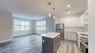 Located at 32 S State St in Concord, NH, this well-designed one-bedroom, one-bathroom unit offers a thoughtful single-floor layout with modern finishes throughout. The open-concept kitchen and living area serves as the heart of the space, featuring a two-tone center island with a crisp white countertop and pendant lighting above. The kitchen is fully equipped with stainless steel appliances — including a refrigerator, range, microwave, and dishwasher — complemented by white shaker-style cabinetry, a marble-inspired tile backsplash, and ample upper and lower storage. The living area flows naturally from the kitchen and is bathed in natural light from multiple large windows that frame views of the surrounding neighborhood. Wood-look plank flooring in a cool gray tone runs consistently throughout the unit, adding warmth and visual continuity. The bedroom is a comfortable size, featuring a double-door closet and additional hallway access. The bathroom is finished with wood-toned cabinetry, a white quartz countertop, a large mirror, and tile-style flooring. Two hallways provide efficient circulation, and a dedicated entrance and closet add to the unit's practical layout. Clean white walls and recessed lighting complete the bright, contemporary aesthetic.