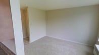 This single-floor space presents a straightforward and functional layout finished in neutral tones throughout. The main living area is carpeted in a light beige, providing a soft underfoot feel, and is framed by smooth white baseboards and clean, understated walls. The space is open and versatile, lending itself well to a variety of furniture arrangements.

An adjacent area, likely a sunroom or secondary living space, features large sliding windows that span much of the wall, allowing a generous amount of natural light to fill the room. These windows offer views of a grassy outdoor area and surrounding buildings, creating a pleasant connection to the exterior. The flooring in this area transitions to a tile surface, adding a practical, easy-to-maintain finish. A wall-mounted air conditioning unit and a baseboard radiator are both present, providing year-round climate control.

A doorway visible in one area hints at additional rooms beyond, suggesting a connected and accessible floor plan. Overall, the space maintains a clean, move-in-ready aesthetic with simple finishes that offer a neutral canvas for personalization.