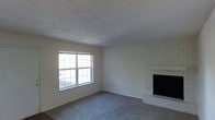 This single-floor residence offers a straightforward and functional layout centered around a welcoming living area. The space features wall-to-wall carpeting in a neutral gray tone, providing a soft and comfortable underfoot feel throughout. A standout feature of the room is the white-painted brick fireplace, positioned in the corner and serving as a classic focal point that adds character and warmth to the interior. Natural light enters through a sizable multi-pane window, brightening the room and creating an open, airy atmosphere. The walls are finished in a clean, neutral tone that complements the overall simplicity of the space, while the textured popcorn ceiling adds a layer of traditional detail overhead. The room's open and uncluttered layout offers flexibility for a variety of furniture arrangements. The overall ambiance is modest and functional, with timeless architectural details like the brick fireplace lending a sense of charm. This space presents a solid foundation with classic features suited for comfortable, everyday living.
