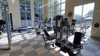 This fitness center offers a bright, open workout environment defined by its soaring floor-to-ceiling windows that line the walls, flooding the space with natural light and providing views of surrounding greenery. The expansive glazing creates an airy, energizing atmosphere that enhances the overall exercise experience.

The facility is outfitted with a comprehensive selection of Life Fitness equipment, including multi-functional cable and weight machines designed for full-body strength training. Cardio equipment, such as stationary bikes and elliptical machines, is positioned to take advantage of the natural light and outdoor views. Free weight benches and additional resistance machines round out the strength training area, providing options for a variety of workout routines.

The flooring features a checkered tile pattern in neutral tones, contributing to the clean, functional aesthetic of the space. Signage indicates the center operates 24 hours, making it accessible at any time of day. The high ceilings amplify the sense of spaciousness, and the layout allows for comfortable movement between stations. Overall, the fitness center combines practical functionality with an inviting, well-lit design.