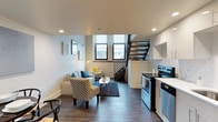 This well-designed two-floor unit offers a thoughtful layout that balances open living with functional detail throughout. The main level features an open-concept floor plan where the living area and kitchen flow seamlessly together, creating a bright and cohesive space. Dark hardwood-style flooring runs throughout, providing a warm contrast to the crisp white walls and recessed lighting overhead.

The kitchen is outfitted with stainless steel appliances — including an electric range, dishwasher, and full-size refrigerator — complemented by white cabinetry, a white subway tile backsplash, and a clean countertop with an undermount sink. Ample upper and lower cabinet storage makes the kitchen as practical as it is visually clean.

The living area offers comfortable gathering space bathed in natural light from large windows that overlook the street below. A sleek metal staircase with black railings connects the main level to the second floor, serving as a striking architectural feature within the open layout.

The bathroom is finished with large-format marble-style wall tiles in the walk-in shower, a modern vanity with a vessel-style sink, and a well-placed mirror. The overall aesthetic across the unit is contemporary and cohesive, with neutral tones, clean lines, and modern fixtures that create an inviting and polished living environment.
