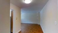 This single-floor space presents a clean, straightforward layout with a quietly classic aesthetic. The most distinctive architectural detail is the rich parquet hardwood flooring, arranged in a traditional herringbone-style pattern that adds warmth and character throughout the main living area. The flooring's warm, honeyed tones contrast elegantly with the bright white walls and white baseboards that frame each room.

The walls are freshly painted in a neutral white, creating a bright and airy atmosphere that feels open and versatile. A flush-mount ceiling light fixture provides even overhead illumination, keeping the space well-lit. The smooth white ceiling complements the overall clean and uncluttered design.

An open doorway is visible to one side, suggesting a connected layout that allows for easy flow between areas. A wall-mounted intercom or switch panel is also present, adding a functional element to the space. The overall ambiance is minimal and move-in ready, offering a blank canvas well-suited for a variety of furnishing styles. The combination of classic parquet floors, neutral walls, and simple fixtures gives this space a timeless quality that balances practicality with understated charm.