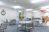 Located at 305 Margaret Ave in Kitchener, Ontario, this single-floor community amenity space offers a well-organized and functional layout designed for social gatherings and recreational activities. The space is divided into two distinct areas, connected by glass-paneled interior doors that allow light to flow between them.

The main room serves as a communal dining and lounge area, furnished with multiple round tables and chairs arranged throughout a carpeted floor. A textured ceiling with flush fluorescent lighting keeps the space bright and welcoming. Along one side, a built-in wooden service counter with a laminate top and tile flooring beneath provides a practical refreshment station, complete with small appliances and a refrigerator. Decorative wall art adds a touch of warmth to the neutral-toned walls.

A dedicated games room adjoins the main area, featuring two dart boards mounted on the wall and ample open floor space, also finished with the same patterned carpet. This room is lit by overhead fluorescent fixtures and offers a casual setting for recreational use.

The space is accessed through a glass-door entrance that connects to a corridor, and an additional interior door provides access to other parts of the building. Overall, the layout is practical and versatile, well-suited for community events, casual dining, and leisure activities.