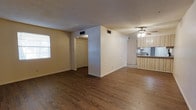 This single-floor space offers a straightforward and functional layout with an open-concept living area that flows seamlessly toward the kitchen. The living room features rich, dark hardwood-style flooring that contrasts cleanly with the light-toned walls, creating a warm and inviting atmosphere. A textured ceiling adds subtle depth to the room, while a ceiling fan with an integrated light fixture provides both comfort and illumination. Natural light filters in through a window on the side wall, brightening the open space. A partial dividing wall separates the living area from the hallway, which leads to additional rooms and a closet door, providing a defined sense of layout and flow. The kitchen is positioned adjacent to the living space and is equipped with light wood cabinetry, offering ample storage. A raised countertop with wood paneling serves as a visual divider between the kitchen and living areas, adding a functional breakfast bar-style element. The kitchen's light-toned flooring contrasts with the darker hardwood in the living room. HVAC vents are visible throughout the ceiling, ensuring climate control across the space. Overall, the unit presents a clean, practical interior well-suited for comfortable everyday living.