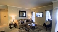 This single-floor space centers around a thoughtfully designed living room that exudes a warm and welcoming ambiance. The room features neutral-toned walls complemented by crown molding, adding a touch of classic elegance to the space. Soft, wall-to-wall carpeting anchors the room and contributes to its cozy feel.

The living area is furnished with a generously sized tan sofa accented with decorative throw pillows in complementary shades of gray and cream. A dark wood coffee table with ornate carved legs serves as the room's centerpiece, positioned atop a patterned area rug that adds visual interest to the floor. A matching end table and table lamp provide ambient lighting on either side of the seating arrangement.

A wall-mounted flat-screen television is positioned for easy viewing, while a large framed mirror on the adjacent wall enhances the sense of space by reflecting natural light. A window fitted with sheer curtains allows natural light to filter into the room, brightening the overall atmosphere. A dark leather accent chair adds contrast and additional seating near the room's edge.