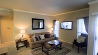 This single-floor space centers around a thoughtfully designed living room that exudes a warm and welcoming ambiance. The room features neutral-toned walls complemented by crown molding, adding a touch of classic elegance to the space. Soft, wall-to-wall carpeting anchors the room and contributes to its cozy feel.

The living area is furnished with a generously sized tan sofa accented with decorative throw pillows in complementary shades of gray and cream. A dark wood coffee table with ornate carved legs serves as the room's centerpiece, positioned atop a patterned area rug that adds visual interest to the floor. A matching end table and table lamp provide ambient lighting on either side of the seating arrangement.

A wall-mounted flat-screen television is positioned for easy viewing, while a large framed mirror on the adjacent wall enhances the sense of space by reflecting natural light. A window fitted with sheer curtains allows natural light to filter into the room, brightening the overall atmosphere. A dark leather accent chair adds contrast and additional seating near the room's edge.