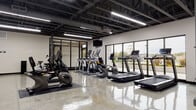This single-floor fitness facility presents a modern industrial aesthetic defined by an exposed steel truss ceiling finished in matte black, polished concrete floors that reflect the abundant overhead linear lighting, and expansive floor-to-ceiling windows along one wall that invite natural light and views of the surrounding outdoor greenery. The open layout provides a generous, unobstructed workout environment divided into distinct functional zones. The cardio area is well-equipped with treadmills, elliptical trainers, stationary bikes, and a rowing machine, all positioned to take advantage of the natural light streaming through the large windows. A dedicated strength training zone features a cable machine, adjustable bench, dumbbell rack, and kettlebells arranged on protective rubber flooring. A separate open floor area accommodates functional fitness activities, with exercise mats, stability balls, and resistance accessories stored neatly on a freestanding rack. Wall-mounted televisions are positioned throughout the space. Exposed ductwork and structural steel columns reinforce the industrial character, while the clean white walls and high ceilings contribute to an airy, motivating atmosphere throughout.