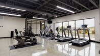 This single-floor fitness facility presents a modern industrial aesthetic defined by an exposed steel truss ceiling finished in matte black, polished concrete floors that reflect the abundant overhead linear lighting, and expansive floor-to-ceiling windows along one wall that invite natural light and views of the surrounding outdoor greenery. The open layout provides a generous, unobstructed workout environment divided into distinct functional zones. The cardio area is well-equipped with treadmills, elliptical trainers, stationary bikes, and a rowing machine, all positioned to take advantage of the natural light streaming through the large windows. A dedicated strength training zone features a cable machine, adjustable bench, dumbbell rack, and kettlebells arranged on protective rubber flooring. A separate open floor area accommodates functional fitness activities, with exercise mats, stability balls, and resistance accessories stored neatly on a freestanding rack. Wall-mounted televisions are positioned throughout the space. Exposed ductwork and structural steel columns reinforce the industrial character, while the clean white walls and high ceilings contribute to an airy, motivating atmosphere throughout.
