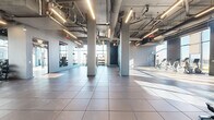 Located in Atlanta, GA (30308), this large-scale fitness center delivers an impressive workout environment defined by its industrial-modern aesthetic and generous open floor plan. The space spans a single level, offering an expansive, column-punctuated layout that accommodates a wide variety of fitness zones without feeling congested.

The architectural design features exposed ductwork and structural elements overhead, complemented by sleek linear LED lighting suspended from the high ceilings. Large floor-to-ceiling windows line the perimeter, flooding the interior with abundant natural light and offering views of the surrounding Atlanta cityscape. The flooring throughout combines durable large-format tile with select hardwood sections, delineating different functional areas of the gym.

The strength training zone is well-appointed with cable machine systems, Smith machines, benches, free weights, and a dedicated dumbbell rack. The cardio section houses a row of stationary bikes, ellipticals, and treadmills positioned to take advantage of the natural light and city views. Open floor space provides room for functional training and group exercise. Exposed brick accent walls add warmth and texture, contrasting with the otherwise sleek, gray-toned palette. Strategically placed mirrors enhance spatial depth throughout the facility.