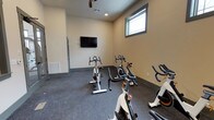 This single-floor fitness space is purpose-built for indoor cycling and cardio exercise. The room features multiple stationary cycling bikes arranged to face a wall-mounted flat-screen television, allowing users to follow workout programs or entertainment during sessions. The layout is straightforward and functional, maximizing the available floor space for equipment use.

The interior is finished with neutral beige walls and durable dark speckled flooring, typical of gym environments designed to withstand heavy use. Recessed lighting is complemented by a ceiling fan, which aids in air circulation during intense workouts. A window positioned high on one wall allows a modest amount of natural light to enter the space without compromising privacy.

Ventilation is further supported by a wall-mounted HVAC vent near the entry door, ensuring comfortable climate control during exercise. The entry door appears to be a standard interior door with a gray frame, providing easy access. Overall, the space offers a clean, utilitarian environment suited for group or individual cycling sessions, with minimal distractions and practical amenities to support an effective workout experience.