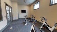 This single-floor fitness space is purpose-built for indoor cycling and cardio exercise. The room features multiple stationary cycling bikes arranged to face a wall-mounted flat-screen television, allowing users to follow workout programs or entertainment during sessions. The layout is straightforward and functional, maximizing the available floor space for equipment use.

The interior is finished with neutral beige walls and durable dark speckled flooring, typical of gym environments designed to withstand heavy use. Recessed lighting is complemented by a ceiling fan, which aids in air circulation during intense workouts. A window positioned high on one wall allows a modest amount of natural light to enter the space without compromising privacy.

Ventilation is further supported by a wall-mounted HVAC vent near the entry door, ensuring comfortable climate control during exercise. The entry door appears to be a standard interior door with a gray frame, providing easy access. Overall, the space offers a clean, utilitarian environment suited for group or individual cycling sessions, with minimal distractions and practical amenities to support an effective workout experience.