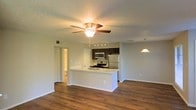 This single-floor space offers a bright and functional open-concept layout that seamlessly connects the kitchen and living areas. The kitchen serves as the centerpiece, featuring a peninsula with a clean white countertop that doubles as a breakfast bar, dark cabinetry, and a full suite of stainless steel appliances including a refrigerator, range, and over-the-range microwave. A pendant light hangs nearby, adding a warm accent to the space. Rich hardwood flooring extends throughout, lending warmth and visual continuity across the entire floor plan. The living area is generously proportioned, illuminated by a ceiling fan with an integrated light fixture that provides both comfort and ambiance. A textured ceiling adds subtle architectural character overhead. Neutral, light-toned walls create a clean and versatile backdrop throughout. A hallway visible from the main living space leads to additional rooms, with a glimpse of a bathroom beyond. Recessed track lighting in the kitchen enhances task visibility, while the overall layout promotes an airy, well-connected feel that is both practical and inviting.