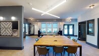 This single-floor communal space is thoughtfully designed to accommodate both collaborative work and social gatherings. The layout is anchored by a spacious open area featuring a large, solid wood communal table surrounded by contemporary upholstered chairs, serving as the centerpiece for group meetings or shared work sessions. Overhead, a striking cross-shaped linear pendant light fixture illuminates the space with bright, functional lighting, complementing the natural light that streams in through multiple large windows fitted with roller shades. Built-in window bench seating along the perimeter adds a comfortable, casual element to the room.

The flooring throughout transitions between wood-look tile planks in the main corridor and a geometric herringbone tile pattern in the gathering areas, adding visual interest and defining distinct zones. Dark charcoal-painted columns and accent walls contrast cleanly against the white ceilings and lighter walls, lending the space a modern, polished aesthetic.

A wide, column-lined corridor connects the gathering area to a lounge zone, which features a warm wood-paneled accent wall, bar-height seating, and additional lounge furnishings. Multiple private rooms enclosed with dark-framed glass doors provide quiet, focused workspaces. Wall-mounted sconces, pendant chandeliers, and decorative artwork round out the refined, detail-oriented design of this versatile space.