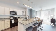 This thoughtfully designed single-floor residence offers a seamless blend of modern aesthetics and functional living. The open-concept kitchen is a standout feature, showcasing granite countertops, a central island with a built-in sink and seating, white cabinetry with open shelving, and a full suite of black appliances including a refrigerator, range, and microwave. Warm hardwood-style flooring runs throughout the kitchen and main living areas, complementing the clean, contemporary palette.

The living area flows naturally from the kitchen, bathed in natural light from large sliding glass doors fitted with vertical blinds that open to an outdoor view. Soft sage-green accent walls add a calming tone to the space.

The bedrooms are comfortably scaled, each featuring plush carpeting, ceiling fans, and generous natural light. One bedroom offers floor-to-ceiling windows with city views, while another includes ample closet space and a dresser area.

Two bathrooms serve the residence with distinction. The primary bath features a dual-sink vanity with granite countertops, large mirrors with sconce lighting, and tile flooring. The secondary bath includes a tub-shower combination with marble-style tile surround and built-in shelving. Together, these spaces deliver a cohesive, polished living environment with modern finishes throughout.