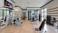 Located at 6925 Roswell Rd in Sandy Springs, Georgia, this fitness center is a well-appointed, single-floor facility designed to support a full range of workout routines. The space features polished concrete flooring throughout, lending it a clean, contemporary aesthetic that is both durable and easy to maintain.

The cardio zone is equipped with multiple treadmills, elliptical machines, and a rowing machine, all positioned to take advantage of the generous natural light streaming in through large, black-framed floor-to-ceiling windows. These windows offer outdoor views of the surrounding greenery and an adjacent pool area, creating an energizing atmosphere during workouts. Multiple wall-mounted flat-screen televisions are positioned throughout the space for entertainment.

The strength training area is equally comprehensive, featuring a variety of cable machines, a multi-station functional trainer, weight benches, and a well-stocked dumbbell rack. A large mirror wall with distinctive horizontal stripe detailing amplifies the sense of space and allows users to monitor their form. The facility also includes direct access to an outdoor area through glass doors, providing convenient indoor-outdoor connectivity. Recessed lighting complements the abundant natural light, ensuring the space is well-lit at all times. The overall ambiance is modern, open, and purpose-built for fitness.