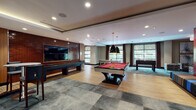 This single-floor amenity space is thoughtfully designed to offer a diverse range of recreational and social experiences. The main game room serves as the centerpiece, featuring a red-felted billiards table illuminated by bold pendant lights, a shuffleboard table set against an exposed brick accent wall with a large flat-screen TV, and a foosball table positioned near floor-to-ceiling windows that flood the area with natural light and views of surrounding greenery. The flooring transitions between hardwood and slate-toned tile, adding visual depth throughout.

Adjacent to the game room is a cozy reading nook distinguished by its rich burnt-orange accent walls, warm wood paneling, and a striking diagonal bookshelf stocked with a wide collection of books. Comfortable, colorfully upholstered seating invites relaxed lounging, while a wall-mounted TV and built-in cabinetry add functional touches to the intimate space.

A connecting corridor with glass-partitioned walls leads to a separate lounge area furnished with tufted ottomans, high-top bistro tables, and bar-height seating—ideal for casual gatherings. The overall design blends modern industrial aesthetics with warm, inviting elements, creating a versatile space suited for both active recreation and quiet relaxation.