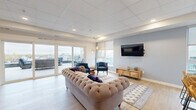 Located at 1530 First Ave N in Moorhead, Minnesota, this community room offers a thoughtfully designed gathering space spread across a single floor. The interior is anchored by a bright, open lounge area finished with light hardwood-style flooring, a drop tile ceiling with recessed lighting, and soft blue-gray accent walls. Large sliding glass doors and generous windows flood the space with natural light while framing views of the surrounding cityscape. The lounge is furnished with a tufted sofa, navy accent chairs, and a wall-mounted TV, creating a comfortable setting for socializing or relaxing. A kitchenette area features warm wood cabinetry, a stainless steel refrigerator, microwave, sink, open shelving, and a large central island with bar seating — ideal for hosting gatherings. A quiet work nook off the main area provides a glass-top desk and curated artwork, offering a focused space for individual use. Sliding doors lead directly to an expansive outdoor terrace equipped with weather-resistant lounge furniture, string lighting, and a partially covered cedar-accented seating area, all surrounded by open skyline views. The terrace is accessible from both sides of the interior, making indoor-outdoor flow seamless and inviting.