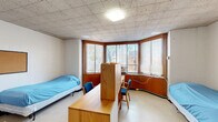 Located at 1825 R Street Northwest in Washington, DC's vibrant Dupont Circle neighborhood, this single-occupancy room offers a functional and efficient layout across one floor. The space is defined by its distinctive curved bay window wall, which spans a wide arc and is fitted with wood-framed windows dressed in horizontal blinds, allowing ample natural light to filter in while offering views of the surrounding urban streetscape. A wall-unit air conditioner is integrated into the window bay, ensuring year-round climate control. The room features warm oak-toned built-in cabinetry along the window wall, providing generous storage and a dedicated desk area. Additional furnishings include a dresser with a wall-mounted shelf and round mirror, offering practical everyday storage. The entrance area leads directly into the main room, with two closets conveniently positioned to maximize organization. The ceiling features a grid tile system with flush lighting fixtures, and the light-toned vinyl tile flooring runs throughout, contributing to a clean, utilitarian aesthetic. White painted block walls lend a straightforward, institutional character to the space. The room's thoughtful layout and built-in features make it a well-equipped and self-contained living space in the heart of Washington, DC.