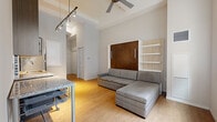This thoughtfully designed studio makes excellent use of its compact footprint through smart layout choices and multifunctional furnishings. The open-plan main living area is anchored by warm hardwood-style flooring that runs throughout, complemented by soaring ceilings that lend an airy, expansive feel to the space. A ceiling fan with integrated lighting enhances comfort, while adjustable track lighting adds a modern, flexible illumination solution.

The kitchenette is well-appointed with dark wood cabinetry, granite countertops, glass-front upper cabinets, a mosaic tile backsplash, stainless steel appliances including a full-sized refrigerator and microwave, and a sink — all efficiently organized along one wall. A window in the living area brings in natural light, brightening the open space.

A built-in Murphy bed system with rich wood paneling serves as the room's focal point, cleverly integrating shelving units on either side to maximize storage without sacrificing style. The hallway connects to a closet with louvered doors, offering practical storage, and leads to the entry door.

The bathroom is clean and modern, featuring a white vanity with an undermount sink, a framed mirror, hardwood-style flooring, and a full bathtub with tiled surround — a complete, functional retreat within the studio's efficient design.