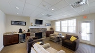 This single-floor communal lounge is designed to provide a comfortable and inviting environment for relaxation and socializing. The room's centerpiece is a floor-to-ceiling stacked stone fireplace topped with a white mantel, above which a wall-mounted flat-screen television is positioned for easy viewing. Recessed lighting embedded in a drop-panel ceiling ensures the space is well-lit throughout.

The seating arrangement includes a mix of plush sofas and upholstered armchairs in neutral earth tones, complemented by small round side tables that offer convenient surface space. A yellow accent pillow adds a subtle pop of color to the otherwise warm, understated palette. Carpeted flooring throughout contributes to the room's cozy atmosphere.

Along one wall, a beverage station with a countertop, cabinetry, and a small under-counter refrigerator provides convenient refreshment access. Large windows along the adjacent wall allow natural light to fill the space, enhancing its open and airy feel. Dark wood cabinetry flanks the fireplace, adding a refined touch to the overall design. The space presents a practical yet comfortable setting suited for everyday use.