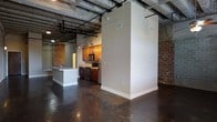 This single-floor loft blends industrial character with modern functionality, creating a distinctive living environment. The space is defined by its exposed brick walls, which lend warmth and texture throughout, while the polished concrete floors provide a sleek, durable foundation that reflects the loft's urban aesthetic. Overhead, an open ceiling reveals structural beams and exposed ductwork, adding to the industrial charm and contributing to a sense of generous vertical space.

The kitchen is thoughtfully appointed, featuring warm-toned wood cabinetry paired with stainless steel appliances, including a refrigerator, microwave, and range. A central kitchen island with a white finish offers additional prep and casual dining space. Track lighting above the kitchen ensures the area is well-lit and practical.

A structural column and partial partition wall define the transition between the kitchen and the open living area, maintaining a sense of flow while subtly zoning the space. The open-concept layout allows for flexible furniture arrangement throughout the living area. Together, these elements create a cohesive, loft-style environment that balances raw architectural details with contemporary finishes.