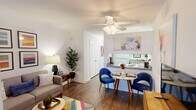 This single-story studio in Metairie, Louisiana, offers a thoughtfully organized open-concept layout that maximizes every square foot. The main living area features dark hardwood-style flooring throughout, crisp white walls, and a ceiling fan with integrated lighting, contributing to a bright and airy atmosphere. A built-in shelving column serves as a subtle architectural divider between the living and sleeping areas, providing both storage and visual separation without fully enclosing the space.

The living area accommodates a comfortable seating arrangement alongside a media console, while a colorful area rug adds a vibrant focal point underfoot. A gallery wall of bold, colorful framed artwork enhances the cheerful character of the space. The kitchen features a pass-through counter with granite-style surfaces that opens directly into the dining area, creating a seamless connection between cooking and entertaining. The dining nook comfortably fits a small bistro-style table and chairs.

The sleeping area, partially defined by the shelving unit, offers a cozy and intimate feel. A hallway connects the main living space to the full bathroom, which includes a combination tub and shower with overhead cabinetry for storage. White panel doors and neutral wall tones throughout lend the studio a clean, cohesive aesthetic.