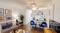 This single-story studio in Metairie, Louisiana, offers a thoughtfully organized open-concept layout that maximizes every square foot. The main living area features dark hardwood-style flooring throughout, crisp white walls, and a ceiling fan with integrated lighting, contributing to a bright and airy atmosphere. A built-in shelving column serves as a subtle architectural divider between the living and sleeping areas, providing both storage and visual separation without fully enclosing the space.

The living area accommodates a comfortable seating arrangement alongside a media console, while a colorful area rug adds a vibrant focal point underfoot. A gallery wall of bold, colorful framed artwork enhances the cheerful character of the space. The kitchen features a pass-through counter with granite-style surfaces that opens directly into the dining area, creating a seamless connection between cooking and entertaining. The dining nook comfortably fits a small bistro-style table and chairs.

The sleeping area, partially defined by the shelving unit, offers a cozy and intimate feel. A hallway connects the main living space to the full bathroom, which includes a combination tub and shower with overhead cabinetry for storage. White panel doors and neutral wall tones throughout lend the studio a clean, cohesive aesthetic.