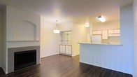 This single-floor residence offers an open-concept layout that seamlessly connects the living, dining, and kitchen areas into one cohesive and functional space. Rich, dark hardwood flooring extends throughout, providing a warm and grounded aesthetic that complements the bright, neutral-toned walls.

The living area is anchored by a classic fireplace with a clean tile surround, adding both charm and a focal point to the room. A decorative arched niche accents the wall nearby, lending an architectural touch to the space. A stylish pendant light fixture hangs centrally, illuminating the dining area with an elegant glow.

A built-in shelving unit with glass shelves and a wooden base serves as a tasteful divider between the living and dining areas, offering display space while maintaining an open feel throughout.

The kitchen features a peninsula-style counter that defines the cooking zone while keeping it visually connected to the rest of the living space. White cabinetry and a full-size refrigerator provide ample storage and functionality. Additional upper cabinetry offers generous storage capacity.

Overall, the space presents a clean, well-maintained interior with a balanced mix of practical features and timeless design elements, making it a comfortable and inviting single-floor home.