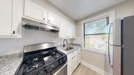 This thoughtfully designed 2-bedroom, 1-bathroom unit offers a clean, modern aesthetic with quality finishes throughout its single floor. The layout is connected by multiple hallways, providing a practical flow between rooms. The kitchen is a standout feature, showcasing white shaker-style cabinetry with black hardware, granite countertops with subtle grey veining, a subway tile backsplash, and stainless steel appliances including a gas range and full-size refrigerator. A window above the sink allows natural light to brighten the space while offering outdoor views of lush greenery. The spacious primary bedroom features wood-tone luxury vinyl plank flooring, neutral wall tones, and two windows that fill the room with natural light. The second bedroom is equally bright, with a window framing a green outdoor view and matching flooring throughout. The bathroom is elegantly finished with large-format marble-look wall tiles surrounding a full soaking tub and shower combination, accented by brushed gold fixtures and a natural light-filled window. Consistent vinyl plank flooring, neutral warm-grey walls, and crisp white trim unify the entire unit, creating a cohesive and inviting atmosphere. A dedicated closet provides additional storage.