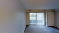 This single-floor space offers a clean, functional layout designed for comfortable everyday living. The main living area is a standout feature, highlighted by a full-width sliding glass door dressed with vertical blinds that filter natural light while offering a pleasant view of the surrounding greenery outdoors. The room is finished with soft, wall-to-wall carpeting underfoot, contributing to a warm and cozy atmosphere. Neutral-toned walls keep the space feeling open and versatile, providing an easy backdrop for a variety of interior styles. Ceiling vents are positioned throughout, indicating a centralized HVAC system for year-round climate control. The overall design is straightforward and practical, with clean lines and a modest aesthetic that prioritizes livability. The abundance of natural light sourced through the large glass door gives the interior a bright, airy feel despite its understated finishes. This space is well-suited for those seeking a low-maintenance, move-in ready environment with a connection to the outdoors through its generous glazing and garden-facing outlook.
