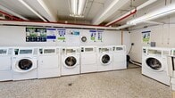 Located at 3435 Rue Drummond in Montréal, this shared laundry facility is a practical, well-organized space comprising a main laundry room and a connecting hallway. The interior is finished with white tile walls and durable terrazzo flooring throughout, lending the space a clean, utilitarian character. Overhead fluorescent lighting ensures the area is brightly and evenly illuminated, complemented by an exposed ceiling with visible piping and ventilation infrastructure.

The main laundry room is equipped with a row of commercial-grade front-loading washing machines and dryers, providing ample capacity for shared use. A deep utility sink station with a stainless steel basin offers additional functionality for hand-washing or pre-treating garments. An Axelius payment terminal is conveniently mounted near the folding counter, enabling card-based or digital payment for machine use. Instructional and informational notices are posted on the walls in both French and English, reflecting the bilingual nature of the Montréal community.

The hallway area features a built-in white countertop that serves as a dedicated folding and sorting station, with a waste bin positioned below for convenience. A wooden bench provides a comfortable waiting area for residents. The space is designed with functionality and ease of use in mind, making it a well-equipped shared amenity for building residents.