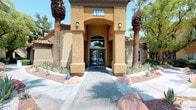 This single-story building presents a striking exterior characterized by warm stucco façades, arched entryways, and a terra cotta tile roofline, framed by desert landscaping with native plants, decorative boulders, and tall palm trees. A wide, welcoming walkway leads to the glass-paneled entry doors beneath a bold covered portico.

Inside, the space opens into a soaring, voluminous interior that immediately draws attention upward toward a dramatic sculptural chandelier with branching arms and exposed Edison-style bulbs. The exceptionally high ceilings amplify the sense of openness throughout. The main area is thoughtfully arranged with a comfortable seating zone featuring contemporary grey upholstered chairs accented with navy throw pillows, positioned around a glass accent table. A herringbone-patterned tile floor adds visual texture and refinement underfoot.

A central wooden communal table with modern Eames-style chairs serves as a collaborative or meeting area, positioned in front of a mounted flat-screen television and a stylish tiled feature wall with decorative shelving. Large arched windows allow abundant natural light to flood the interior. Beyond the main room, a glass partition reveals an adjacent fitness or amenity space, enhancing the functional, community-oriented character of the overall layout.