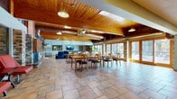 This single-floor clubhouse is defined by its warm, lodge-inspired architecture. Tongue-and-groove wood-paneled ceilings span the main gathering areas, complemented by exposed cylindrical ductwork, pendant lighting, and ceiling fans that reinforce the rustic-industrial character of the space. Brick columns punctuate the interior, adding structural definition while contributing to the overall aesthetic cohesion.

The primary gathering area is generously proportioned, accommodating multiple large wooden dining tables with upholstered chairs, making it well-suited for community events and group gatherings. Large sliding glass doors and wood-framed French doors line the exterior walls, flooding the space with natural light and providing direct access to an outdoor patio furnished with seating and a grill.

A separate lounge zone features leather seating arranged around a wall-mounted television, offering a more casual setting. Vivid mural artwork on one wall adds a bold visual accent near the entrance corridor. A secondary seating nook with wicker chairs sits adjacent to a window overlooking a fitness room.

The kitchen is equipped with stainless steel appliances, including a gas range and full-size refrigerator, granite countertops, and light wood cabinetry, all set against a teal accent wall. Throughout, stone tile flooring unifies each zone, creating a cohesive and functional community space.