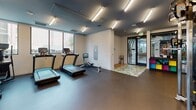 Located at 635 Robinson Ave in San Diego's Hillcrest neighborhood, this fitness center is a well-appointed, single-floor facility designed to support a comprehensive workout experience. The space is divided into distinct functional zones, each dedicated to a specific type of training. The cardio area features multiple treadmills, elliptical machines, and a rowing machine, all positioned near large windows that allow natural light to fill the space and offer views of the surrounding neighborhood. The strength training zone is extensively equipped with a full dumbbell rack, kettlebells, medicine balls, a punching bag, cable machines, benches, and free weight stations, providing everything needed for a thorough resistance training session. Mirrored walls expand the visual depth of the space and allow users to monitor their form. The flooring throughout consists of durable rubber matting suited for high-impact activity. Overhead, a dark ceiling accented by rows of bright linear fluorescent fixtures gives the space a modern, focused aesthetic. Glass partition walls and framed glass entry doors connect the zones fluidly, maintaining an open and airy feel. The overall design is clean, functional, and well-organized, making this fitness center a highly practical and inviting environment for exercise.