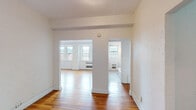 This single-floor unit presents a well-organized layout with multiple living spaces, all unified by warm hardwood flooring and a crisp white color palette. The floor plan, as seen in the 3D model, reveals a thoughtful arrangement of rooms that flow naturally from one to the next.

The spacious living area is particularly notable, featuring multiple gridded windows that flood the space with natural light and offer views of the surrounding neighborhood. Crown molding details and a fireplace mantle add classic architectural character to this central gathering space. An adjacent room, suitable as a formal dining or secondary living area, maintains the same bright aesthetic with additional windows and a radiator unit.

The bedrooms are well-proportioned with hardwood floors and generous windows. One bedroom benefits from a radiator heater and a door providing additional access. The kitchen is functionally equipped with warm wood cabinetry, a stainless steel sink, dishwasher, and electric range, plus a window providing natural light.

Two bathrooms serve the unit: one features classic black-and-white mosaic tile flooring, white subway tile walls, and a soaking tub, while the second offers a pedestal sink and shower. Both maintain a timeless, clean aesthetic consistent throughout the space.