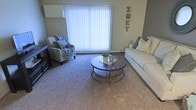 The living room serves as the central gathering area of this single-story space, finished with wall-to-wall carpet in a neutral tone that adds warmth underfoot. A large window dressed with vertical blinds dominates one wall, allowing an abundance of natural light to fill the room while offering adjustable privacy. The space accommodates a white sofa accented with decorative pillows and a patterned armchair, both positioned around a circular two-tiered wooden coffee table with a metallic frame, creating a comfortable and functional seating arrangement. A dark entertainment unit anchors one corner of the room. The walls are painted in a muted gray tone, providing a versatile backdrop that complements the neutral furnishings throughout. Decorative lettering on the wall adds a personalized touch to the overall aesthetic. The layout feels open and well-proportioned, offering a relaxed ambiance suited for everyday living. The combination of soft carpeting, generous natural light, and thoughtfully arranged furniture contributes to an inviting and practical living environment.
