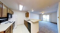This single-floor space is designed with an open-concept layout that seamlessly connects the kitchen and living area. The kitchen features warm wood-toned cabinetry, a double basin sink, a black dishwasher, and a white refrigerator. Light-colored countertops extend into a peninsula-style breakfast bar, providing additional workspace and a casual dining option while maintaining a visual connection between the kitchen and the adjoining living space.

The kitchen is finished with light vinyl tile flooring and is illuminated by a skylight-style flush ceiling fixture, ensuring a bright and functional work environment. A track light fixture adds further task lighting.

The living area transitions to plush carpeting, creating a comfortable and defined zone within the open floor plan. A ceiling fan with an integrated light fixture provides both air circulation and ambient lighting. A sliding glass door floods the space with natural light and offers direct access to an outdoor area, enhancing the overall sense of openness. The neutral white walls throughout create a clean, versatile backdrop that complements the space's practical design.