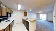 This single-floor space is designed with an open-concept layout that seamlessly connects the kitchen and living area. The kitchen features warm wood-toned cabinetry, a double basin sink, a black dishwasher, and a white refrigerator. Light-colored countertops extend into a peninsula-style breakfast bar, providing additional workspace and a casual dining option while maintaining a visual connection between the kitchen and the adjoining living space.

The kitchen is finished with light vinyl tile flooring and is illuminated by a skylight-style flush ceiling fixture, ensuring a bright and functional work environment. A track light fixture adds further task lighting.

The living area transitions to plush carpeting, creating a comfortable and defined zone within the open floor plan. A ceiling fan with an integrated light fixture provides both air circulation and ambient lighting. A sliding glass door floods the space with natural light and offers direct access to an outdoor area, enhancing the overall sense of openness. The neutral white walls throughout create a clean, versatile backdrop that complements the space's practical design.