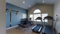 This dedicated fitness space is thoughtfully designed to support a well-rounded workout routine. The room features a vaulted ceiling that lends a sense of openness, complemented by recessed lighting and a striking arched window flanked by two additional windows fitted with white blinds. Together, these windows invite generous natural light while offering views of the surrounding neighborhood.

The space is equipped with a variety of exercise machines, including multiple treadmills and a stationary bike, catering to cardiovascular training needs. A dumbbell rack stocked with a range of free weights is positioned along the blue accent wall, alongside a flat workout bench ideal for strength training exercises. A wall-mounted bracket provides additional storage or support for fitness accessories.

The interior features a neutral-toned floor that is both durable and easy to maintain, while the two-tone wall palette — a deep slate blue on one side and a warm beige on the other — gives the room a modern, energizing aesthetic. The layout is efficient and spacious enough to move freely between equipment. Overall, this fitness room offers a clean, functional, and inviting environment for daily exercise.