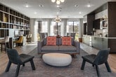 This thoughtfully designed single-floor communal space offers a welcoming and multifunctional environment suited for relaxing, working, and socializing. The open-plan layout is anchored by light wood flooring throughout, creating a warm and cohesive aesthetic that ties the various zones together seamlessly.

The central lounge area features a stylish sofa with accent pillows, paired with contemporary armchairs and a rounded coffee table, all arranged atop a patterned area rug beneath a globe chandelier. Adjacent to this is a dedicated work and study zone, defined by a floor-to-ceiling built-in bookshelf that serves as both a functional library and a striking architectural divider. Bar-height workstations with seating provide space for productivity.

A well-appointed kitchenette area includes cabinetry with dark wood accents, open shelving, a wall-mounted television, a beverage refrigerator, and a sink — complemented by dining tables and teal upholstered chairs, ideal for casual gatherings. Additional lounge seating is distributed throughout, offering flexible arrangements for various group sizes.

Near the main entrance, a grab-and-go market features snack shelving and multiple refrigerated display cases stocked with beverages, providing convenient access to essentials. Large glass entry doors allow natural light to fill the space. The overall ambiance is modern, clean, and inviting, with curated artwork and pops of color adding personality throughout.