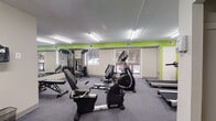 Located at 7 Colony Blvd in Wilmington, Delaware, this fitness center offers a well-organized and fully equipped workout facility on a single floor. The space is designed with functionality in mind, featuring a clean, open layout that accommodates a comprehensive range of exercise equipment. Bold lime-green accent walls contrast with neutral gray tones throughout, creating an energetic and motivating atmosphere. The flooring consists of durable carpet tiles in a gray pattern, providing both comfort and traction during workouts. Overhead fluorescent lighting ensures the space is thoroughly illuminated at all times. A large wall-mounted mirror spans one side of the room, enhancing the sense of space and allowing users to monitor their form during exercise. The cardio section is well-stocked with Spirit-brand treadmills, elliptical trainers, and recumbent stationary bikes, while a multi-station cable machine and adjustable weight bench serve the strength training area. A wall-mounted television is positioned near the treadmills for entertainment during cardio sessions. Windows along one wall allow natural light to filter in, offering views of the surrounding outdoor area. The facility also includes additional rooms accessible via interior doors, adding further utility to the overall space.