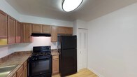 This single-floor space includes a well-equipped kitchen that serves as a central feature of the layout. The kitchen is outfitted with black appliances, including a gas range and a full-sized refrigerator, both by Whirlpool. Warm-toned wood cabinetry lines the upper walls, providing ample storage, while an under-cabinet range hood is installed above the cooktop for ventilation. The countertops feature a dark granite-style finish that contrasts neatly with the lighter cabinetry. Light hardwood flooring runs throughout, adding warmth and continuity to the space. A flush-mount circular ceiling light provides overhead illumination. Adjacent to the kitchen, a louvered bifold door leads to a closet, offering additional storage. The walls are finished in a clean, neutral white, contributing to a bright and open feel. The overall aesthetic is practical and functional, with a cohesive color palette of warm wood tones, black appliances, and white walls that give the space a tidy, move-in-ready appearance.