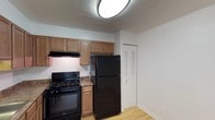 This single-floor space includes a well-equipped kitchen that serves as a central feature of the layout. The kitchen is outfitted with black appliances, including a gas range and a full-sized refrigerator, both by Whirlpool. Warm-toned wood cabinetry lines the upper walls, providing ample storage, while an under-cabinet range hood is installed above the cooktop for ventilation. The countertops feature a dark granite-style finish that contrasts neatly with the lighter cabinetry. Light hardwood flooring runs throughout, adding warmth and continuity to the space. A flush-mount circular ceiling light provides overhead illumination. Adjacent to the kitchen, a louvered bifold door leads to a closet, offering additional storage. The walls are finished in a clean, neutral white, contributing to a bright and open feel. The overall aesthetic is practical and functional, with a cohesive color palette of warm wood tones, black appliances, and white walls that give the space a tidy, move-in-ready appearance.