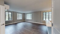 This single-floor unit offers a thoughtfully designed open-concept layout finished with rich, dark hardwood flooring that extends throughout the main living areas. The spacious main living area is bathed in natural light, courtesy of multiple large windows dressed with vertical blinds, all set against neutral grey walls and clean white trim that create a calm, contemporary atmosphere.

The kitchen is a standout feature, showcasing wood-toned cabinetry with modern hardware, marble-patterned countertops, a white tile backsplash, and a full suite of stainless steel appliances including a range, over-the-range microwave, and refrigerator. A peninsula counter separates the kitchen from the living area, adding both functionality and visual definition to the open floor plan.

The bedroom is generously proportioned, offering multiple windows with vertical blinds that let in abundant natural light, along with a set of double closet doors providing ample storage.

The bathroom is cleanly finished with grey tile flooring, white tile wainscoting, a pedestal sink, a large mirror with overhead lighting, a white storage cabinet, and a glass-enclosed shower, completing this well-appointed unit.