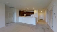 Located at 12001 Old Vine Blvd in Lewes, Delaware, this single-floor residence offers a well-organized and functional layout designed for comfortable everyday living. The open-concept design connects the kitchen and living area seamlessly, creating a spacious and airy feel throughout the main living space. The kitchen is equipped with rich dark wood cabinetry that provides ample storage, light-toned countertops, and white appliances including a built-in microwave and refrigerator. A peninsula-style breakfast bar serves as both a practical workspace and a casual dining area, separating the kitchen from the living space while maintaining an open sightline. Recessed lighting is featured throughout the ceiling, providing consistent and even illumination across the space. The living area is finished with neutral wall-to-wall carpeting, contributing to a warm and inviting atmosphere. A hallway extends from the main living area, leading to additional rooms, each accessible through classic paneled doors that complement the home's clean and understated aesthetic. The overall design emphasizes practicality and ease of movement, making this a well-appointed single-level home in the Lewes, Delaware area.